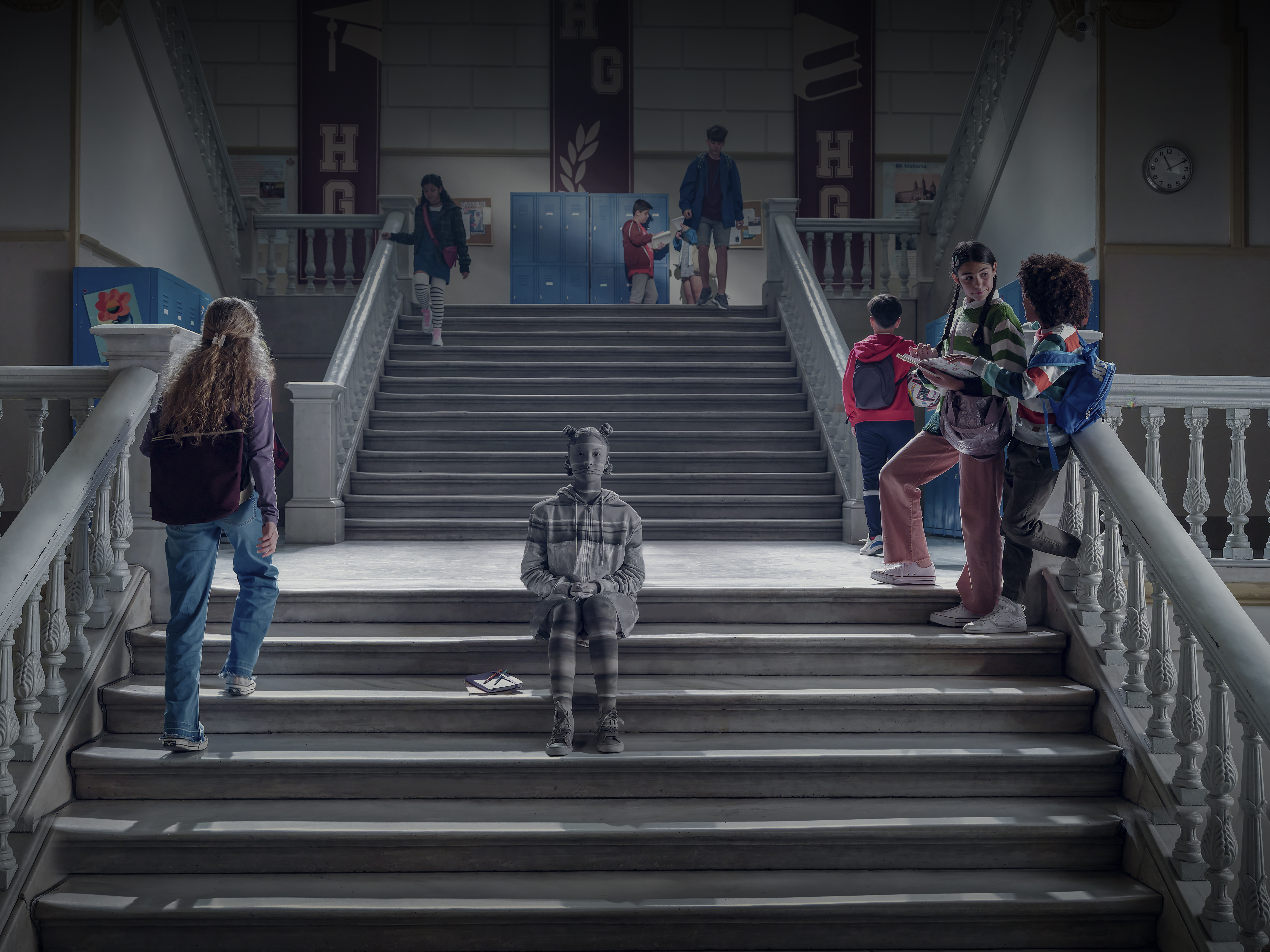 A group of teenagers are standing and walking on the stairs of a high school, with banners in the background and neutral lighting.